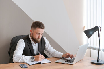 Handsome businessman is working with laptop in office, in business clothes at his desk. A bearded successful guy does his job perfectly well. Reflects and analyzes his actions