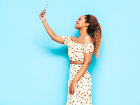 Beautiful black woman with afro curls hairstyle. Smiling model dressed in summer dress. Sexy carefree female posing near blue wall in studio. Tanned and cheerful. Taking selfie