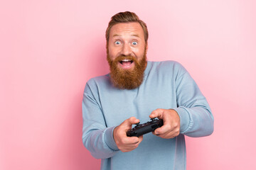 Portrait of handsome guy with red hairstyle wear blue long sleeve hold joystick playing excited game isolated on pink color background