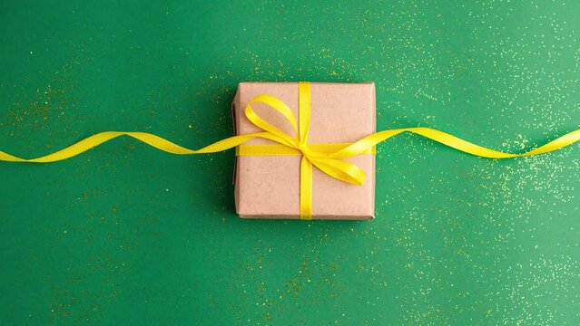 Brown gift box with a yellow satin ribbon bow on jade green background with gold colored glitter. Stop motion animation Christmas Holidays and present concept flat lay.