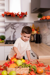 Smiling cute boy standing at counter with fruits, autumn leaves and pumpkin. Autumn decorated kitchen. Holidays concept.