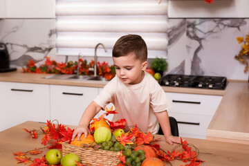 Smiling cute boy standing at counter with fruits, autumn leaves and pumpkin. Autumn decorated kitchen. Holidays concept.