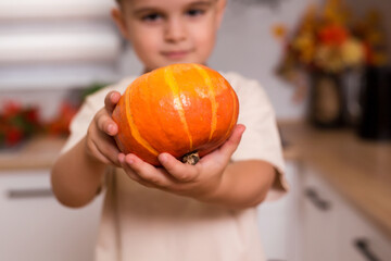Fototapeta premium Caucasian little happy boy holds a fresh pumpkin at kitchen. Healthy lifestyle, diet concept, vegetarianism, raw food and autumn holidays