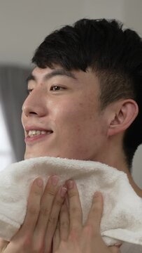 Vertical Screen: happy asian man in undershirt is looking at his reflection in the mirror and wiping face with a towel during daily skincare routine in the morning at home