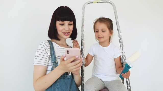 A cheerful happy family is renovating a purchased apartment, mom holding smart phone. The mother hugs her daughter, sitting on a ladder and having video call.