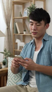 Vertical Screen: leisure Chinese male approaching and sitting down on the comfortable living room sofa to enjoy social media feeds on his smartphone at home during daytime.