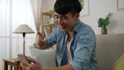 closeup of a carefree asian Korean young man slapping on his lap and having a hearty laugh while watching a funny video on his mobile phone in the living room at home.