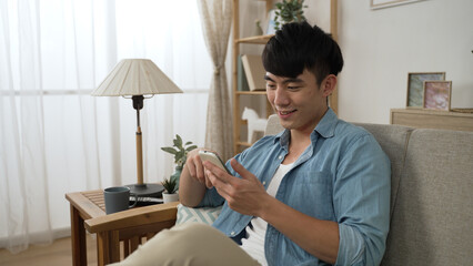 leisure Japanese male approaching and sitting down on the comfortable living room sofa to enjoy social media feeds on his smartphone at home during daytime.