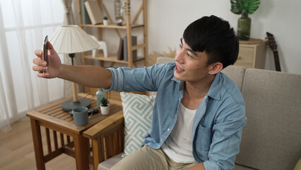 happy asian Korean male is waving hand and finger pointing to mobile phone screen while having fun chatting with friend through video call on the sofa at home