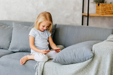 The girl is sitting on the sofa and playing on the phone. The concept of children's addiction to gadgets. Online training. Internet games