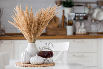Still-life. Pampas dried grass in a vase, white ceramic pumpkins and a teapot on a white table in the interior of a Scandinavian-style home kitchen. Cozy, stylish autumn concept.