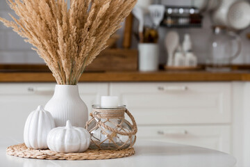 Still-life. Dried pampas grass in a vase, white ceramic pumpkins and a candlestick with a candle on a white table in the interior of a Scandinavian-style home kitchen. Cozy, autumn concept.