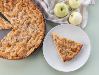 Homemade apple pie on the table in a gray plate with yellow apples and a kitchen towel. A piece of cake in a white plate. Top view banner. Flat background with pastries