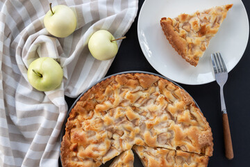 Homemade apple pie on the table in a gray plate with yellow apples and a kitchen towel. A piece of cake in a white plate. Top view banner. Flat background with pastries