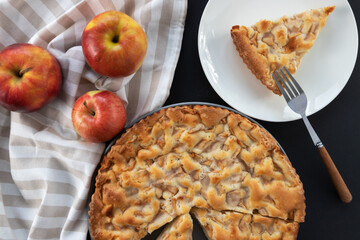 Homemade apple pie on the table in a gray plate with red apples and a kitchen towel. A piece of...
