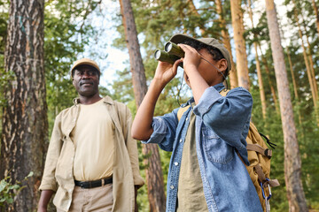 Cute youthful boy looking far away through binoculars while hiking in pine tree forest with his grandfather on summer weekend