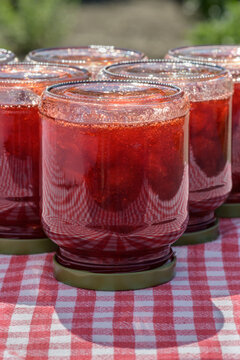 Glass jars with fresh homemade strawberry jam standing upside down on a table with a red checkered tablecloth.
