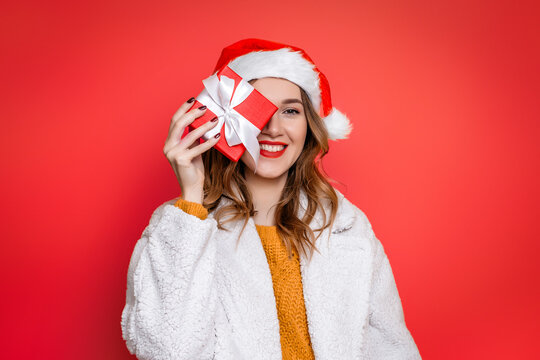 woman in santa hat smiles and covers one eye with a gift box isolated over red background