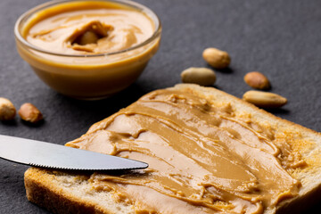 Image of knife, bread slice and bowl with peanut butter on dark grey background
