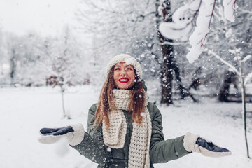 Happy young woman playing with snow in snowy winter park wearing warm knitted clothes and having...