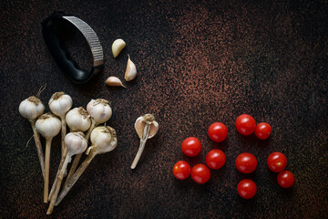 Garlic cloves, cherry tomatoes and garlic press on dark background with copy space