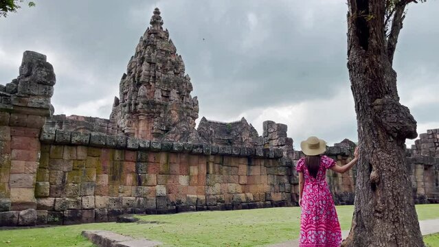 Young tourist woman wearing a red floral dress and a hat stands beside a large tree with the wind blowing in front of Phanom Rung Prasat Hin Phanom Rung. Buriram Province, Thailand