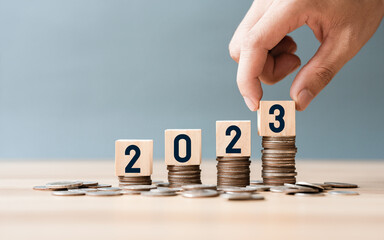 Hand place wooden cube 2023 on top stack of coins with sky blue background and copy space. saving...
