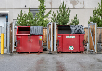 Two red steel industrial garbage and recycling containers in a wooden enclosure in a parking lot after rainfall, daytime, nobody