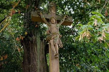 Fototapeta premium Mystische Ort - Altes steinernes Kruzifix unter einem schattigen Baum