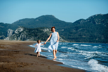 Cute little toddler girl and mother in blue dresses runnig and playing with waves on the wild beach. Turkey, Iztuzu beach,  Dalyan. Mom and daughter
