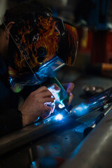 Worker in helmet with flames welding the steel part of a motorbike using an argon welding machine and gloves in a garage