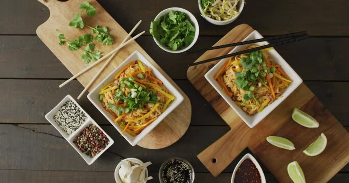 Composition of bowls with pad thai, vegetables, sauces and spices on wooden background