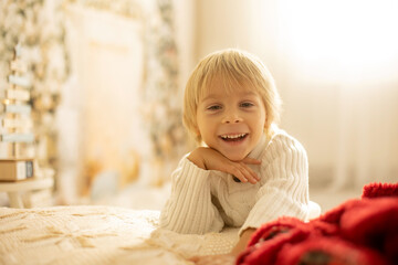 Fototapeta premium Cute blond toddler preschool boy, reading a book and opening present on Christmas on cozy home