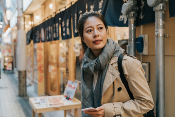 Asian Chinese woman tourist looking at distance while using navigation app on phone to search for directions in the arcade food street in Shinsekai area of Osaka Japan
