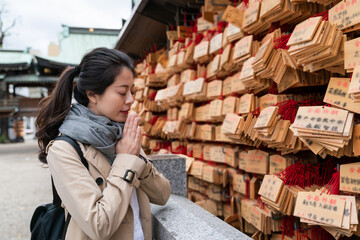 devout Asian Japanese woman worshiper making a wish with hands together by a wall of ema wooden plaques at Osaka Tenmangu Shrine in japan
