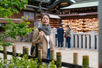 curious Asian Japanese woman visitor looking over bamboo fence into distance while exploring at Osaka Tenmangu Shrine in japan near a shrine with ema wooden luck charms