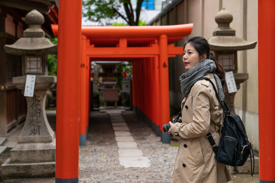Asian Japanese girl photographer turning to look into distance at the entrance of rows of scarlet torii gates while traveling to Osaka Tenmangu Shrine in japan in spring