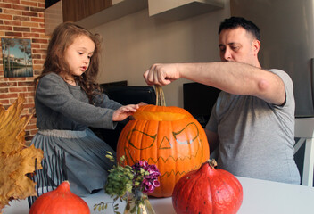 A little girl with long hair and her dad are preparing Jack's Lantern from a large orange pumpkin. The tradition of preparing for the Halloween holiday. Decor of dried leaves, flowers and candles 