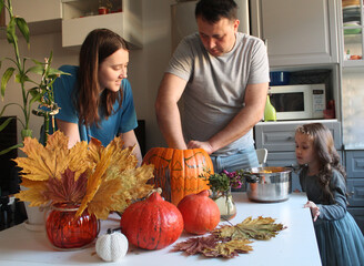 Dad and two daughters make Jack's lantern out of an orange large pumpkin on the background of an autumn decor of dry leaves and candles. Traditions of celebrating Halloween