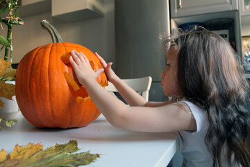A little girl with long hair covers her eyes with her palms to a pumpkin that was made for the Halloween holiday. The tradition of making Jack's lantern for the autumn holiday, All Saints' Day