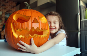 A little girl with long hair hugs a large pumpkin made for the Halloween holiday. The tradition of making Jack's lantern for the autumn holiday, All Saints' Day