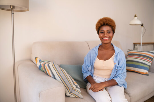 Beautiful black woman in blue shirt sitting in living room on beige sofa attentively looking at camera. Copy space. Light beige colors home. Friendly look. casual relaxed clothes