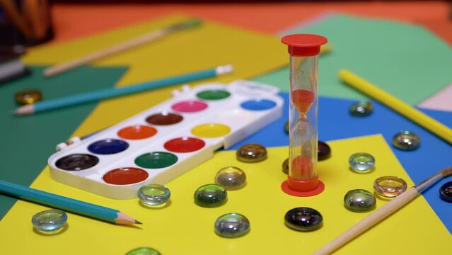 Hourglass on the Background of Scattered Multi Colored Cardboard, School Supplies. Time lapse. Time concept. Flowing sand. End of lesson. Creativity, homework. Yellow background. Childhood. Education.