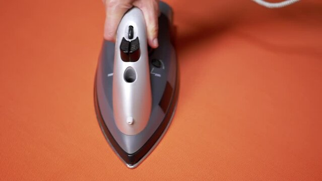 Female Hand Ironing a Bright Orange Cloth with a Steam Iron on an Ironing Board. Close-up. Routine housework. Duties of a hostess. Orange background. Textile. Modern steamer, steam iron. Slow-motion.