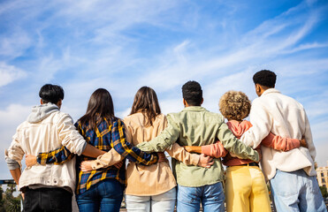 Rear view of young group of happy multiracial friends hugging each other standing in a row looking...