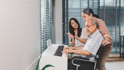 Family home health care wife and daughter caregiver take care senior elderly retirement father at home sitting on the wheel chair playing piano together.