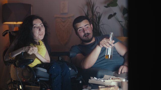 Woman with disability in a wheelchair talking and watching movie or sport games on TV in the evening at home, with her husband who drinking beer