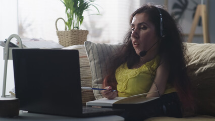 Woman with a disability in headphones sitting on a sofa writing in notebook and talking via video...
