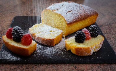 Close-up on the homemade plum cake resting on a black stone cutting board while being dusted with icing sugar. Cooking at home. Delicious healthy homemade dessert served with berries
