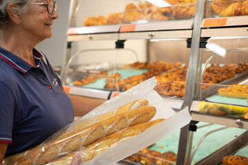Senior caucasian woman in the supermarket holds a paper bag with three baguettes while looking at...
